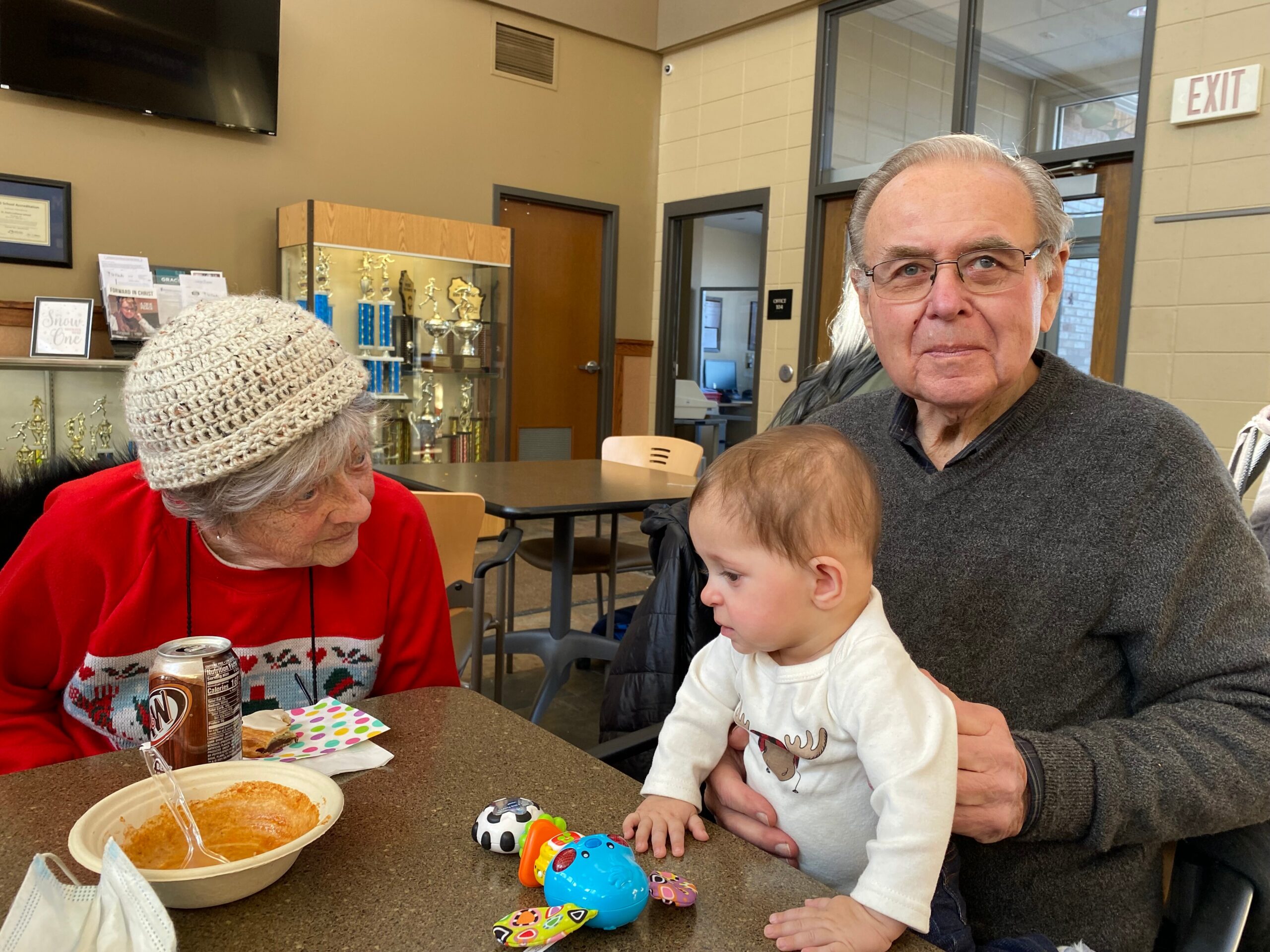 Elderly woman in a red sweater and knit hat sits at a table with soup, while a man holds a baby and a toddler plays with colorful toys nearby in a cafeteria setting.