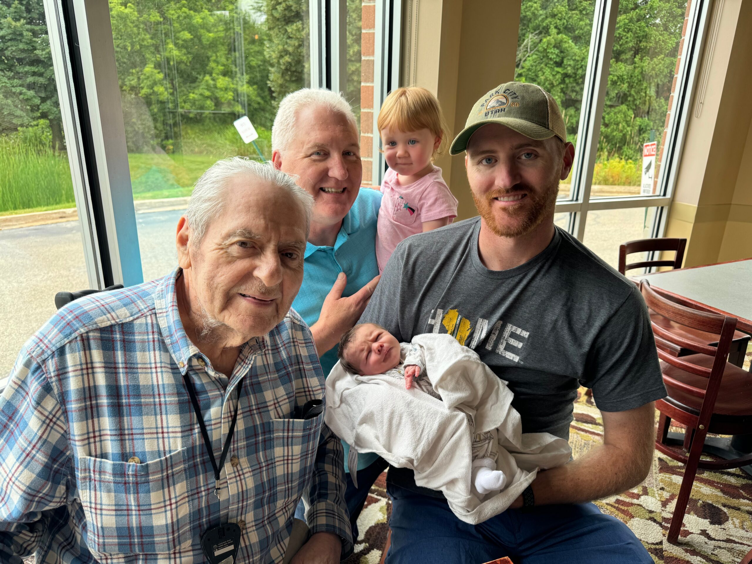 Group portrait indoors with large windows: a smiling man in a blue shirt, an older man in plaid, a young girl, a man in a cap, and a newborn wrapped in white, all posing together.