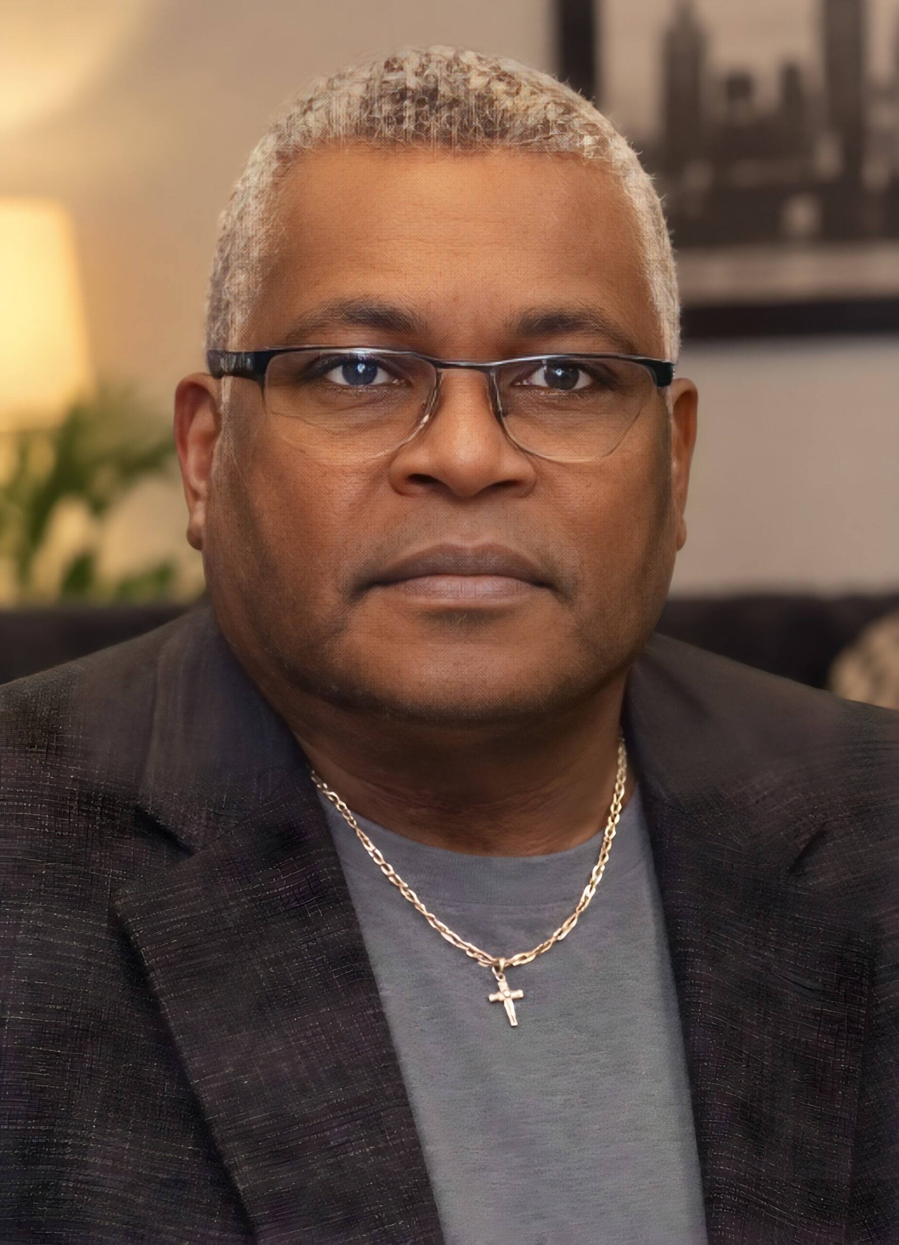 Close-up of a man with short gray hair and glasses wearing a dark blazer and a gold cross necklace.
