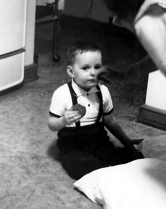Toddler in a white polo shirt and suspenders sits on a carpeted kitchen floor, holding a cookie and looking ahead.