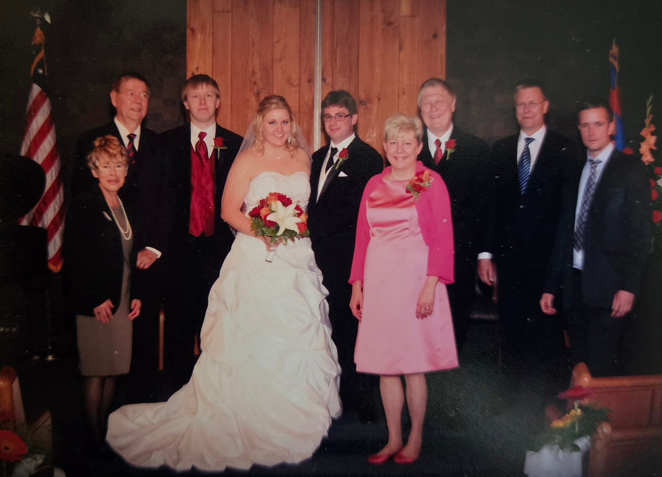 Bride in a strapless white wedding dress holding a bouquet, posing with family on a stage with an American flag nearby.