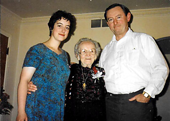 Three people posing together in a living room: a young woman in a blue dress, an elderly woman in black, and a man in a white shirt.