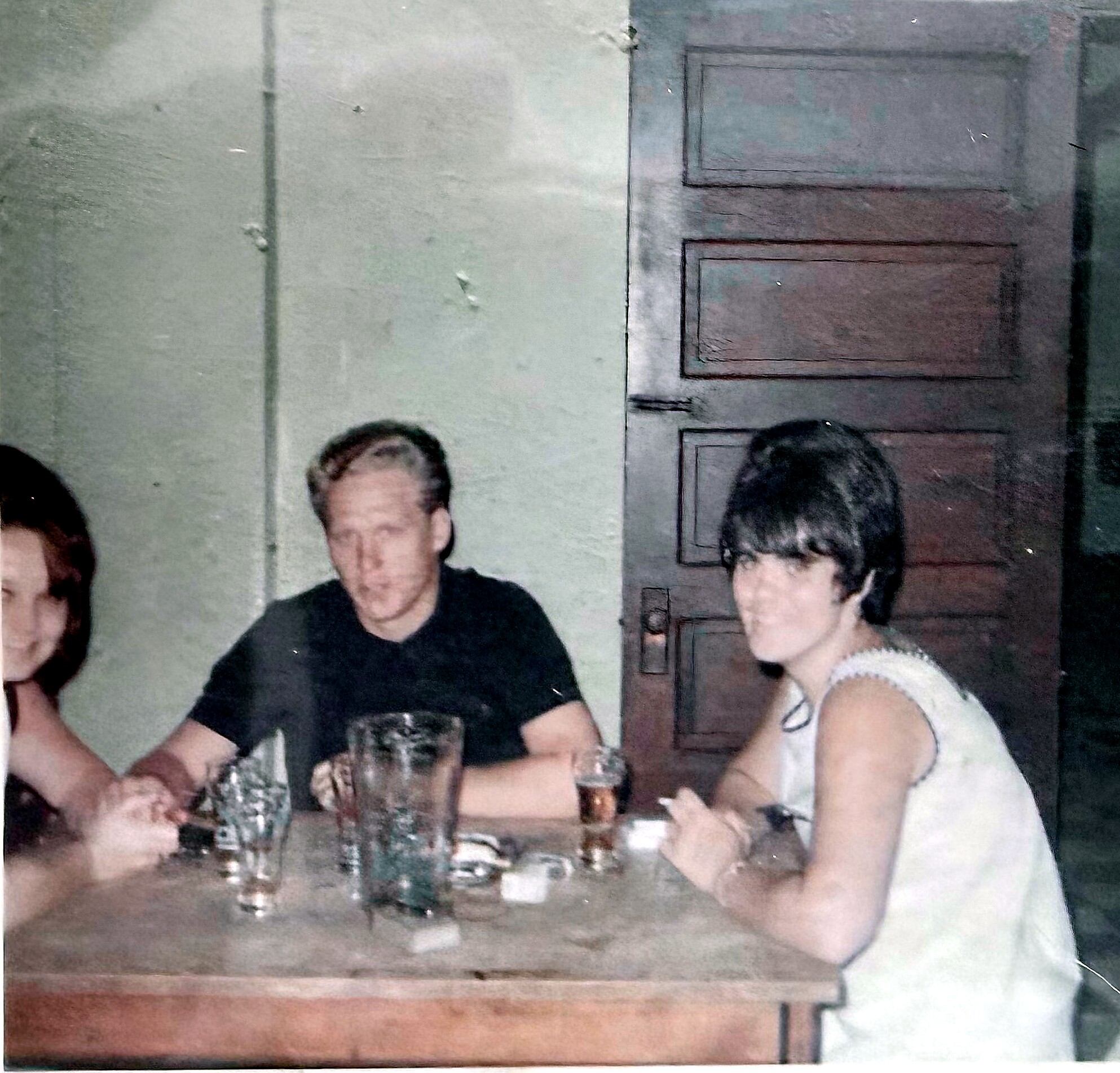 People seated around a table with drinks in a dim room; a wooden door is visible in the background.