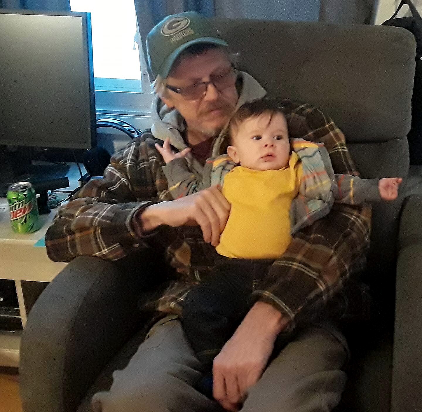 Older man with glasses and a green cap sitting in a chair, cradling a baby in a yellow onesie on his lap indoors.
