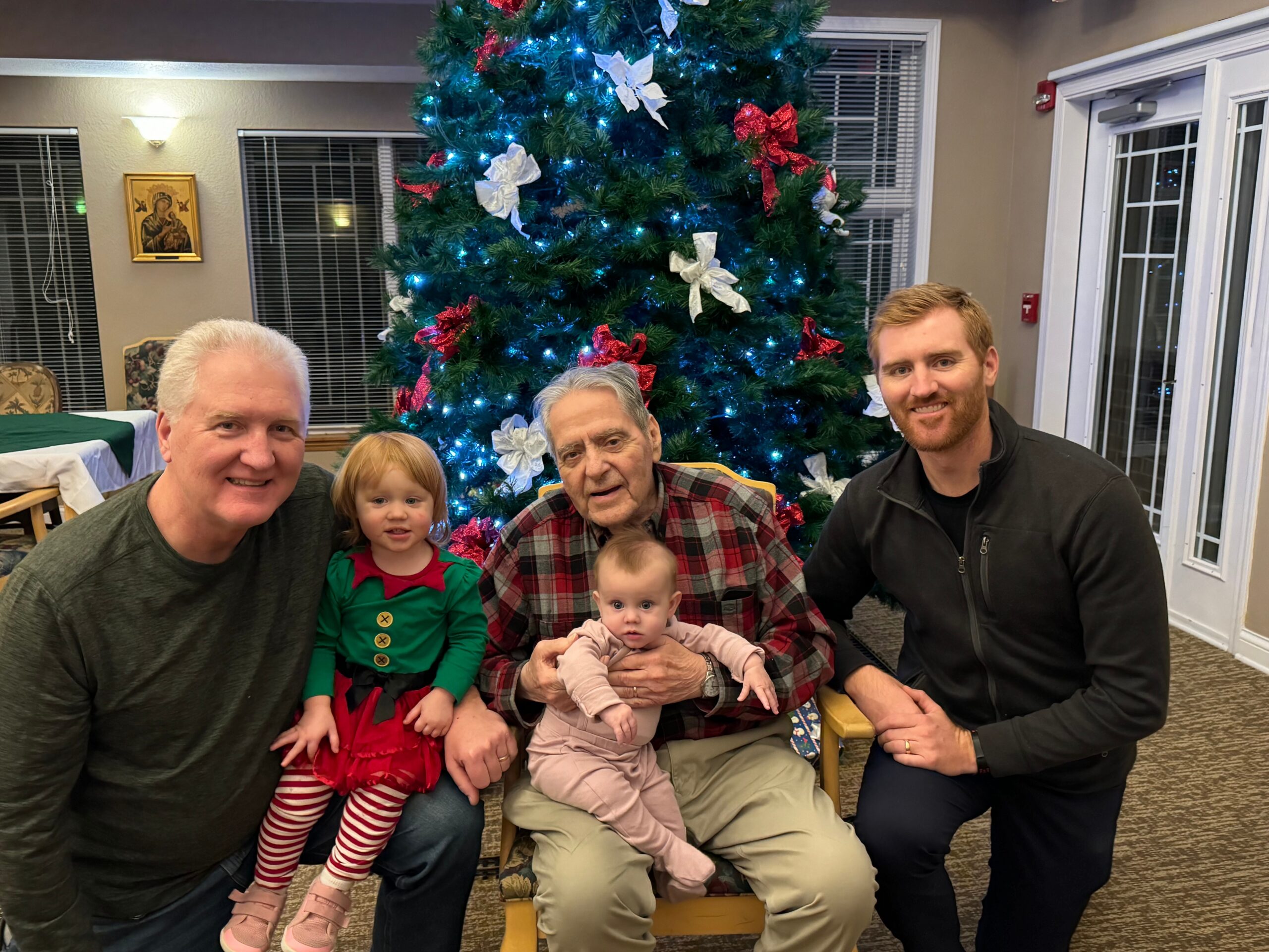 Family group posing indoors in front of a lit, decorated Christmas tree, including two young children in festive outfits and adults smiling at the camera.