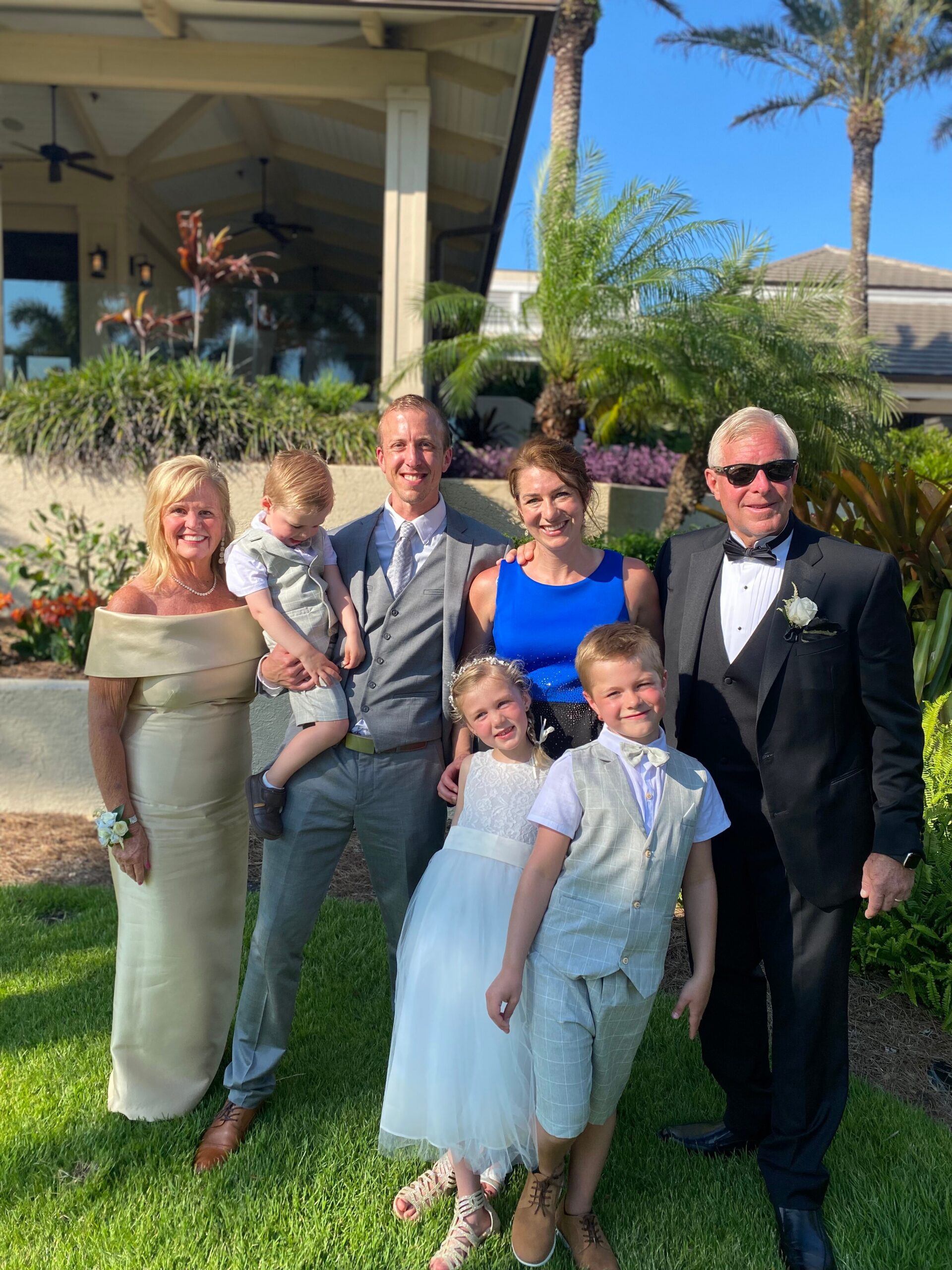 Group of smiling family in formal attire posing on a lawn at an outdoor wedding with palm trees in the background.
