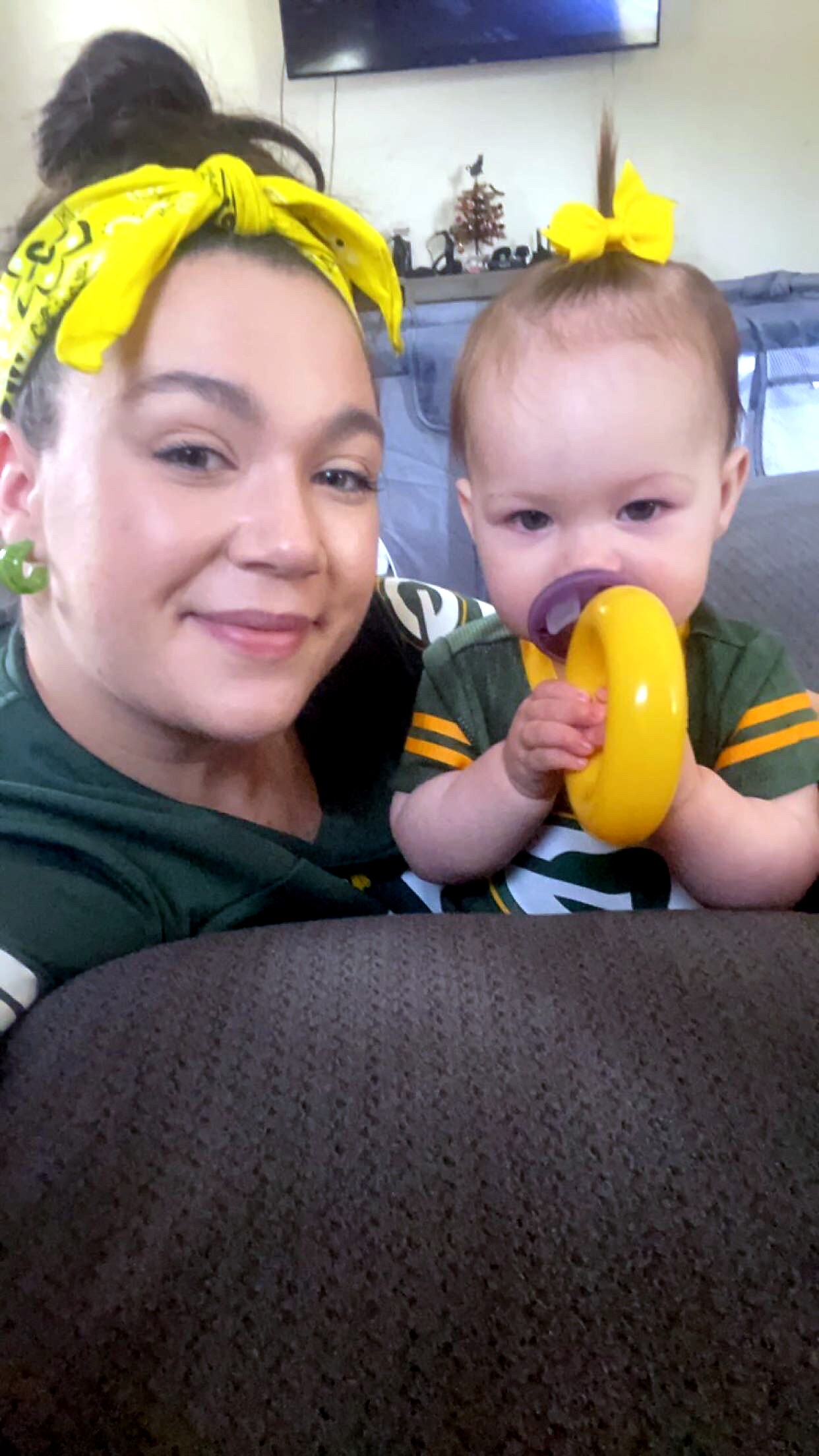 Smiling woman and baby with matching yellow headbands indoors; baby holds a yellow ring toy near their face.