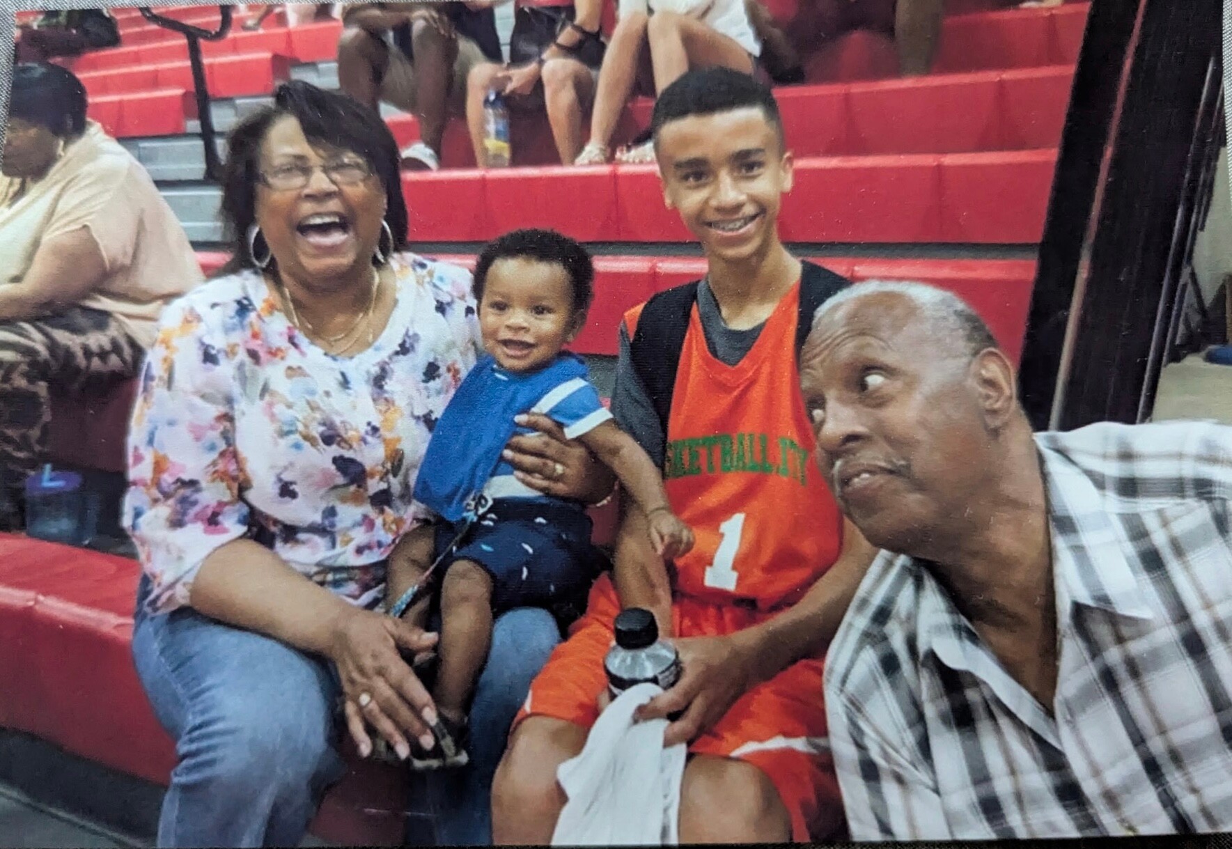 A smiling family posing on red gym bleachers: a woman in a floral blouse holding a baby, a teenage boy in an orange basketball jersey, and a man in a plaid shirt leaning in for a photo.