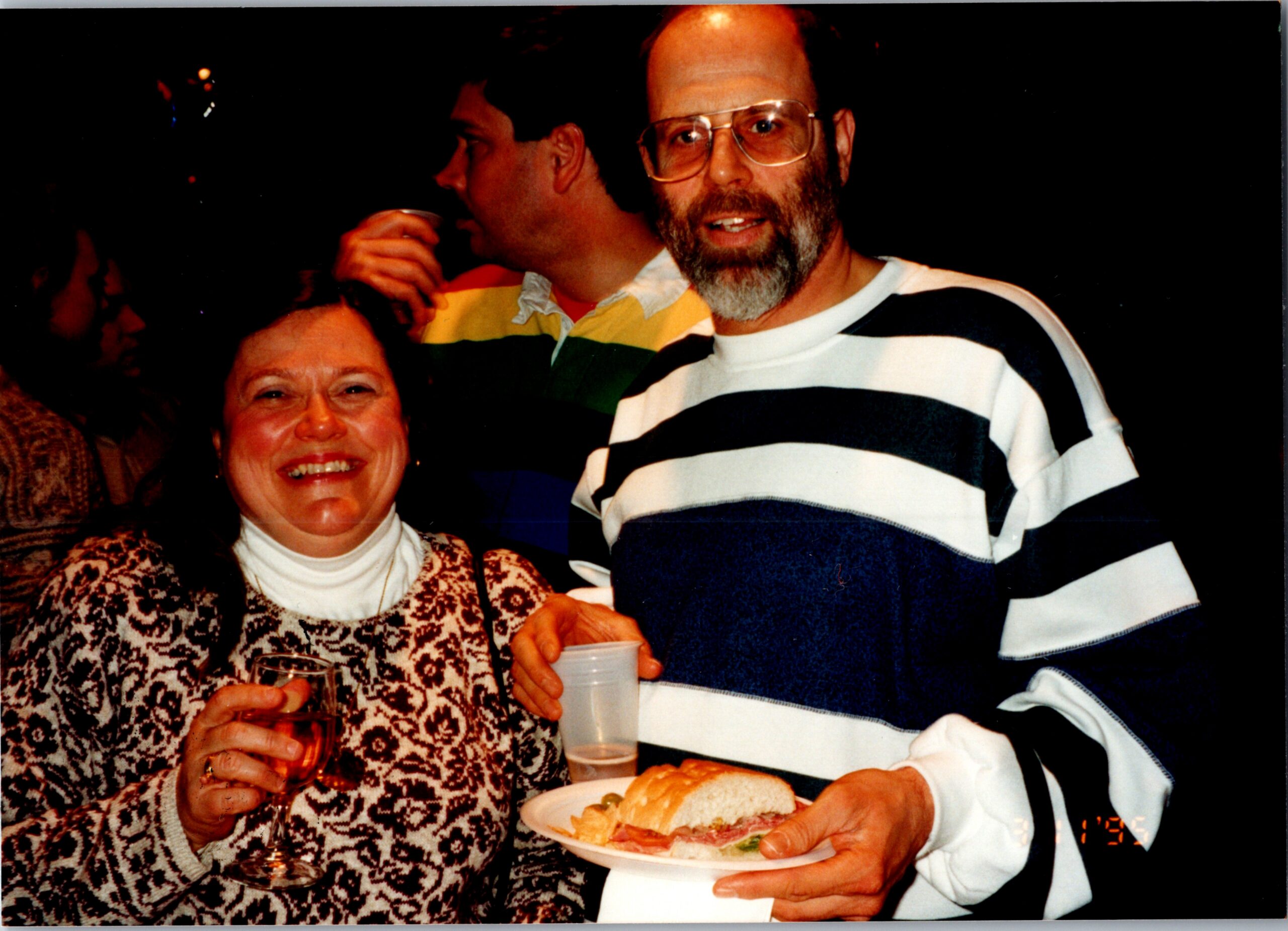 Smiling woman in a patterned sweater holding a glass, standing next to a bearded man in a striped shirt at a social gathering with food in the foreground.