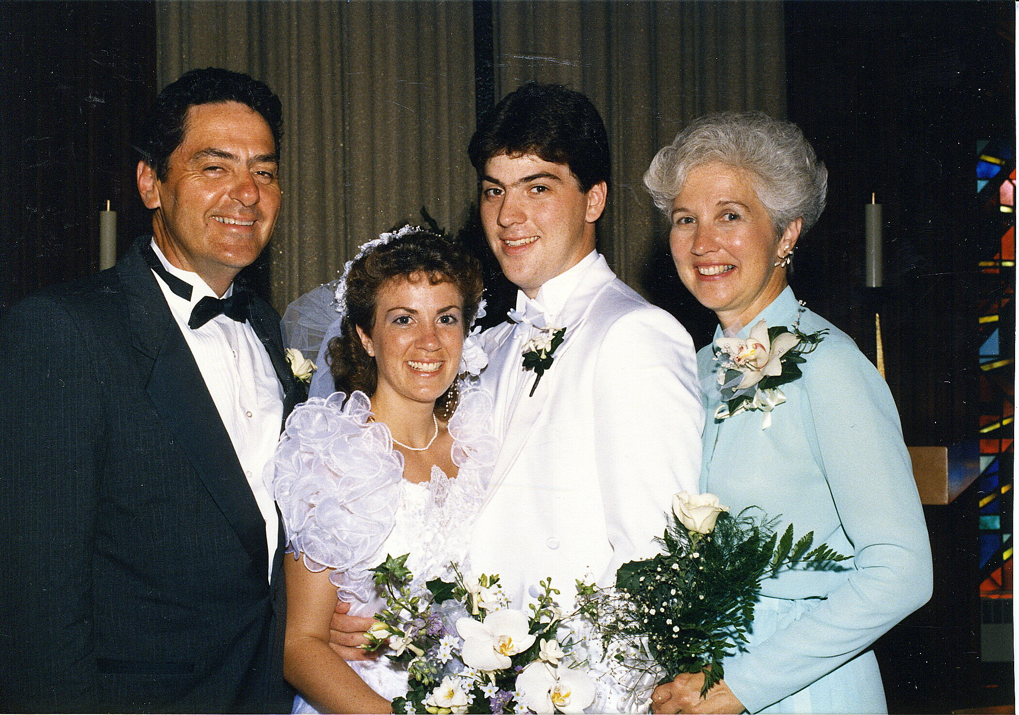 Bride and groom pose with two parents at a wedding Portrait; everyone smiles and holds flowers.