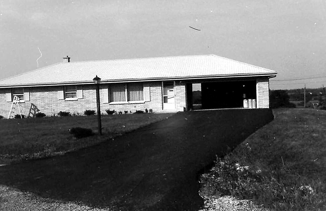 Black-and-white photo of a single-story brick house with a driveway leading to an open carport on the right and a lamp post in the yard.