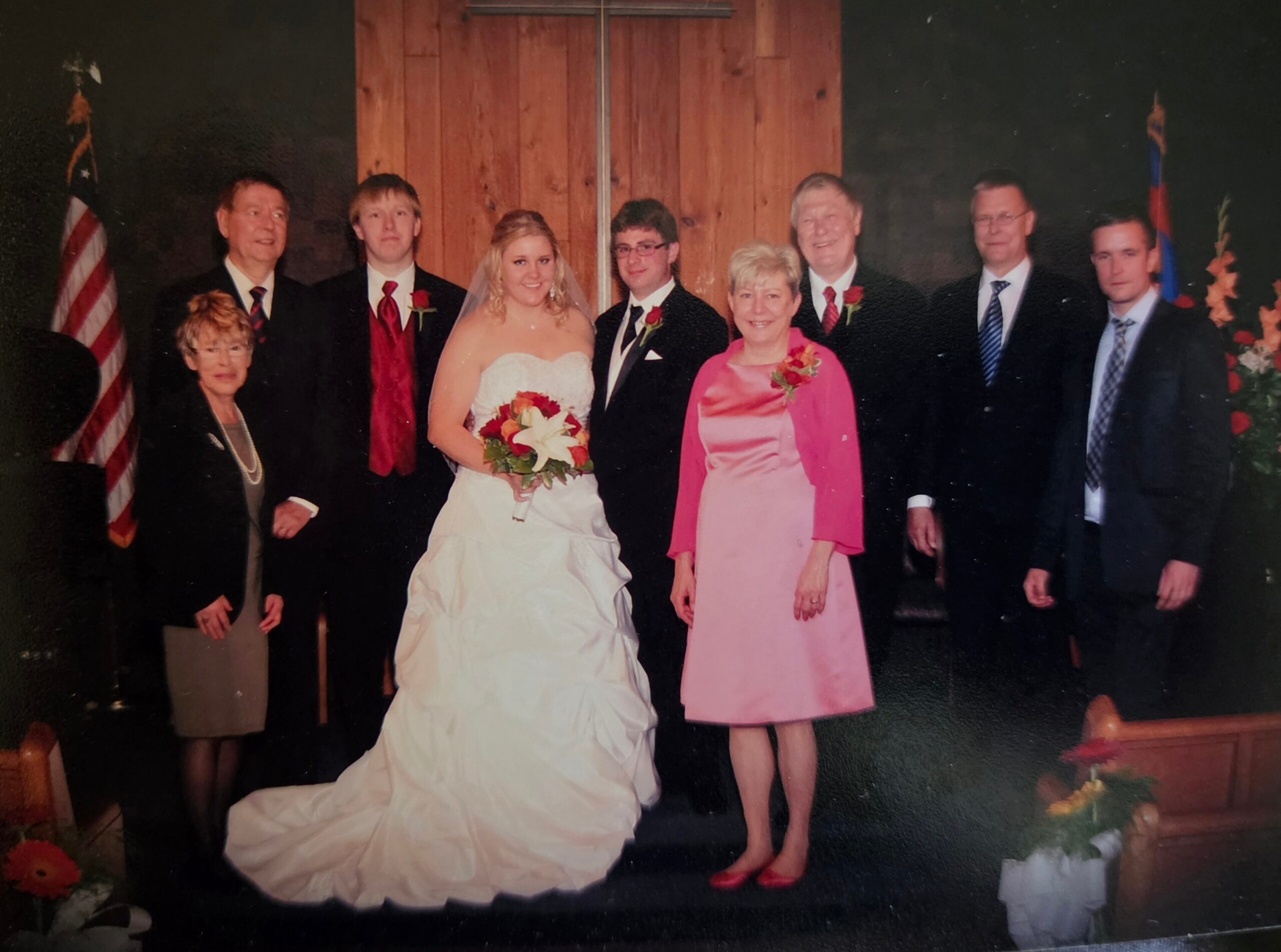Bride in a white wedding gown holding a bouquet, flanked by family in formal attire on a stage with wooden backdrop and flags behind them.