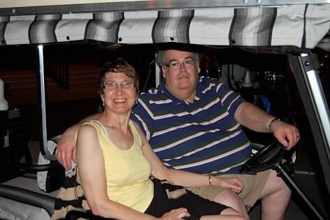 Smiling couple sits close together in a golf cart under a canopy at night; woman wears a yellow sleeveless top, man in a blue striped polo.