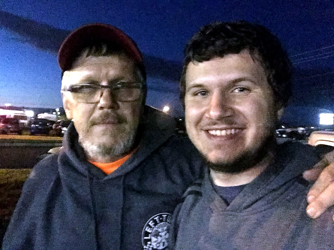 Two men take a nighttime selfie, smiling at the camera, outdoors in a parking lot.