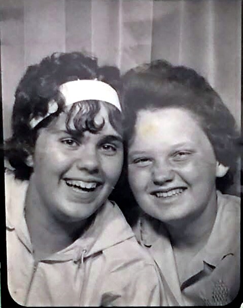 Two women smiling together in a vintage black-and-white photo, one wearing a white headband beneath curls.