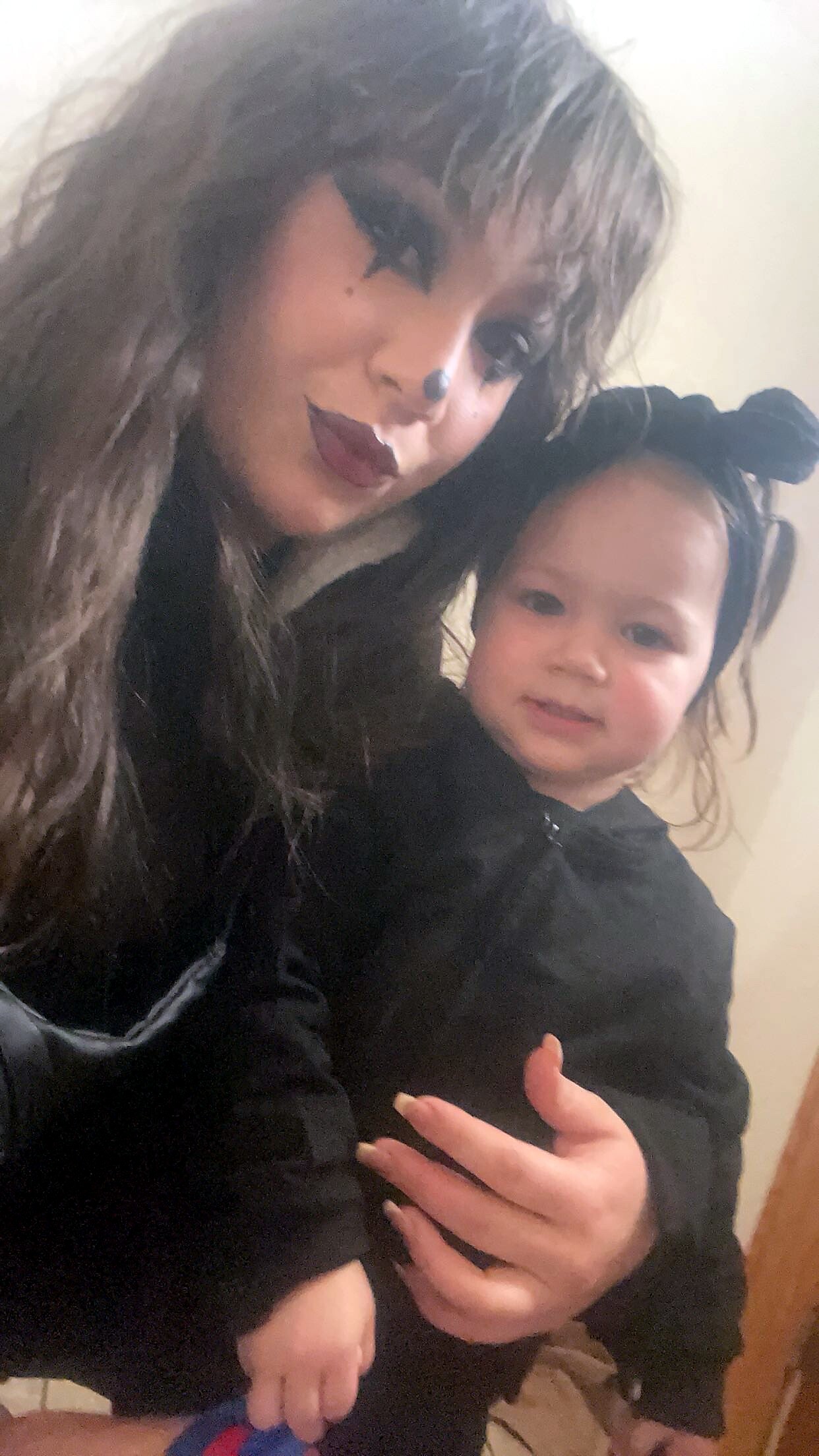 Adult wearing a long dark wig and clown makeup holding a smiling toddler in a black outfit indoors.