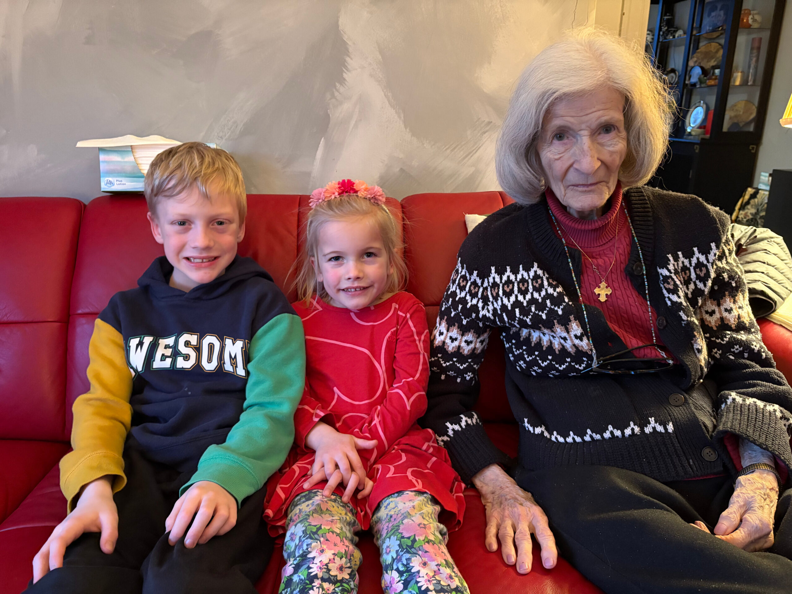 Three generations sitting on a red couch: a boy, a girl in red, and an elderly woman with gray hair wearing a black and white sweater accessorized with a necklace.