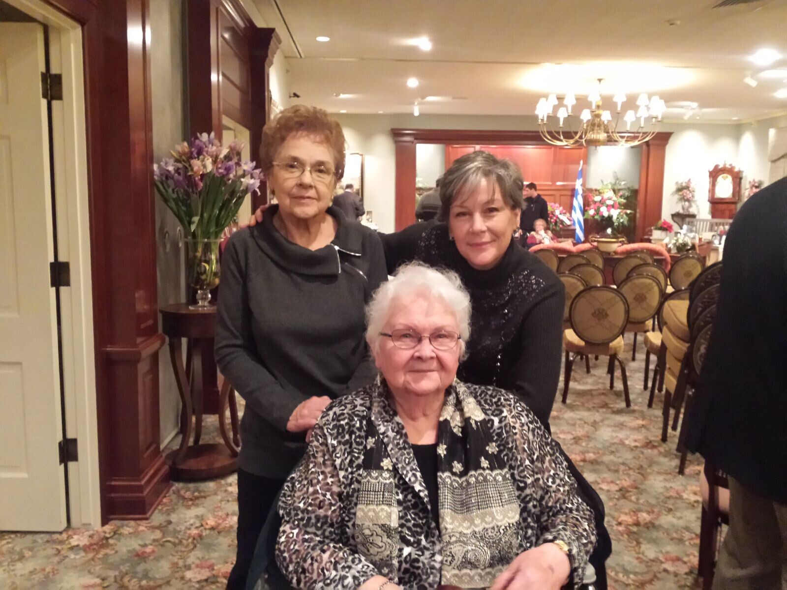 Three women pose together in a decorated room; one seated in front wearing a patterned blouse while two stand behind her, smiling at the camera.