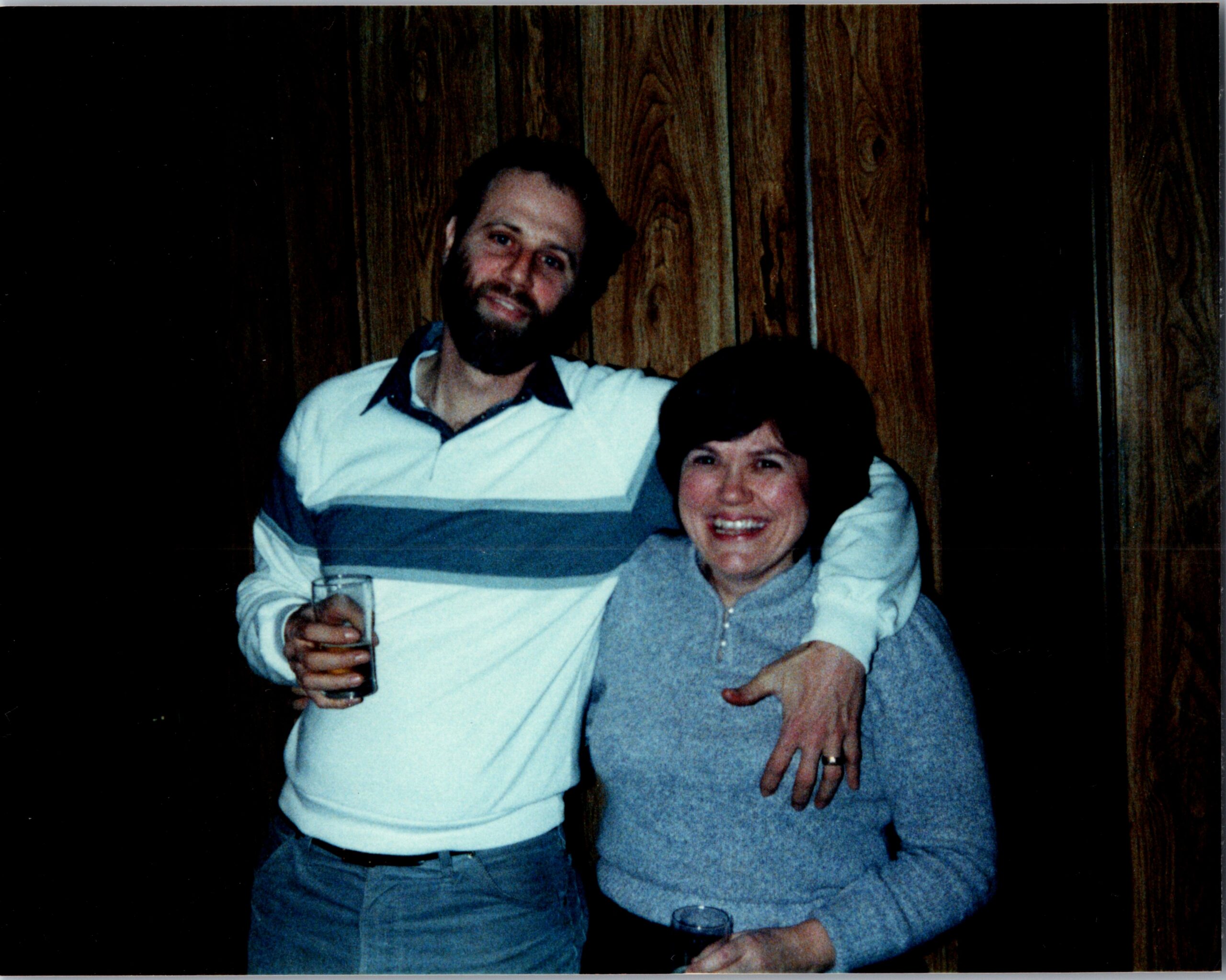 Two friends posing for a photo indoors, the man holding a glass and wearing a white and gray striped shirt beside a smiling woman in a gray sweater against a wood-paneled wall.