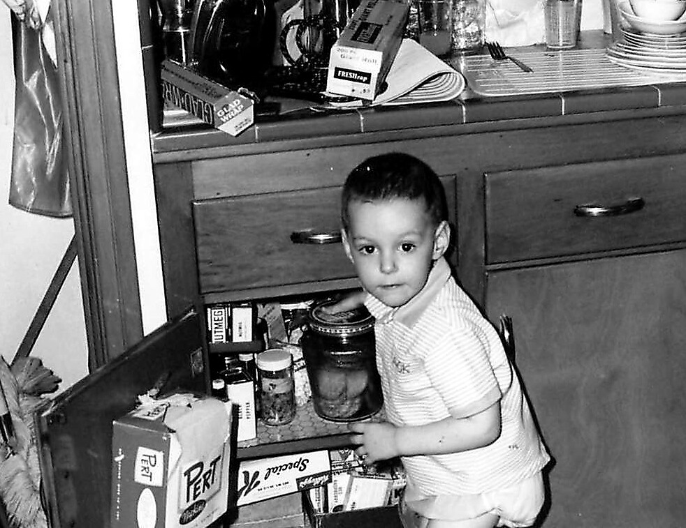 Young child in a striped polo standing beside an open under-cabinet in the kitchen, reaching into shelves with jars and boxes visible in the cabinet.