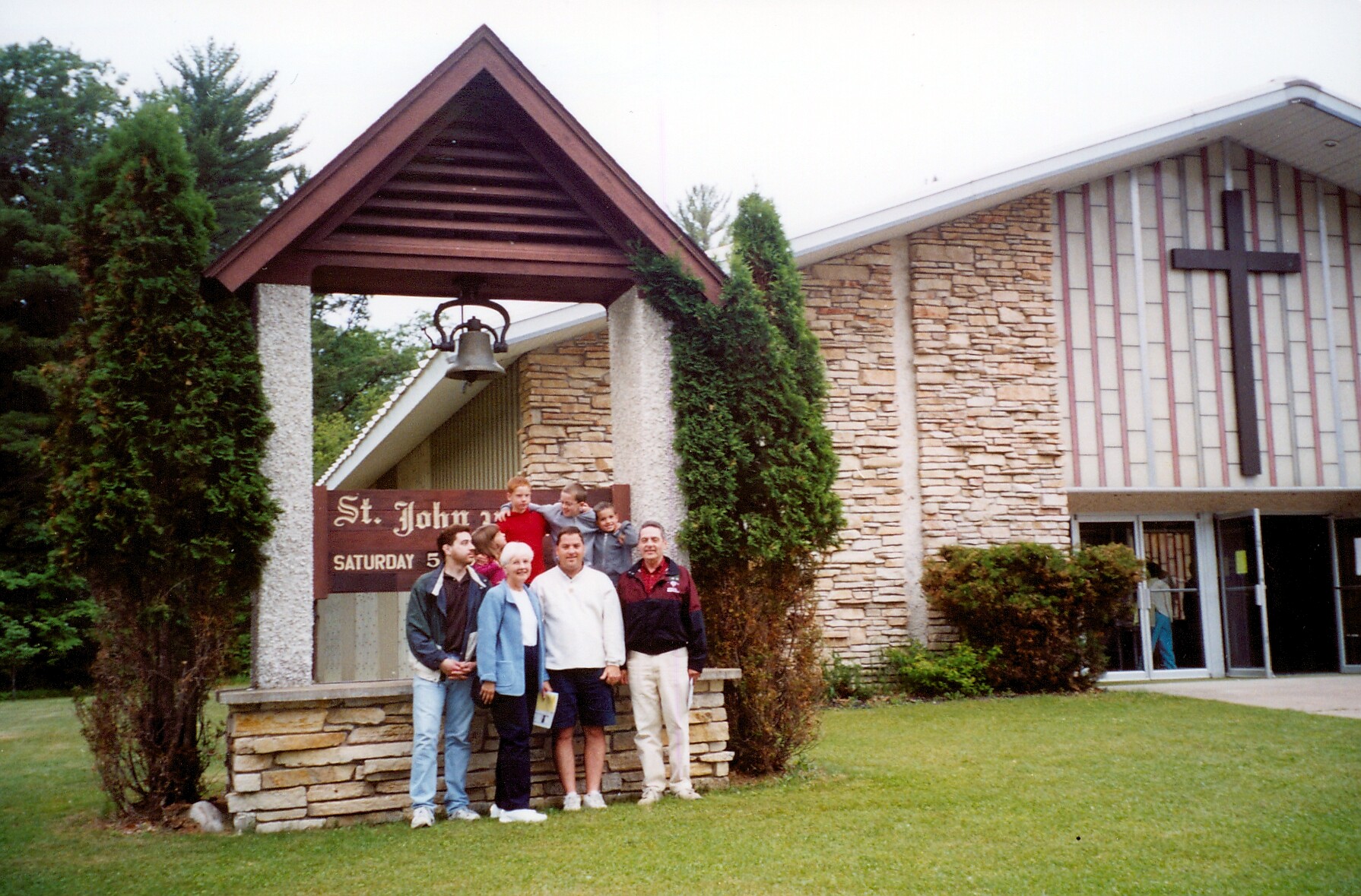 Group of nine people posing together in front of St. John church entrance, with a stone façade, cross on the wall, and a wooden sign reading 'St. John' nearby.