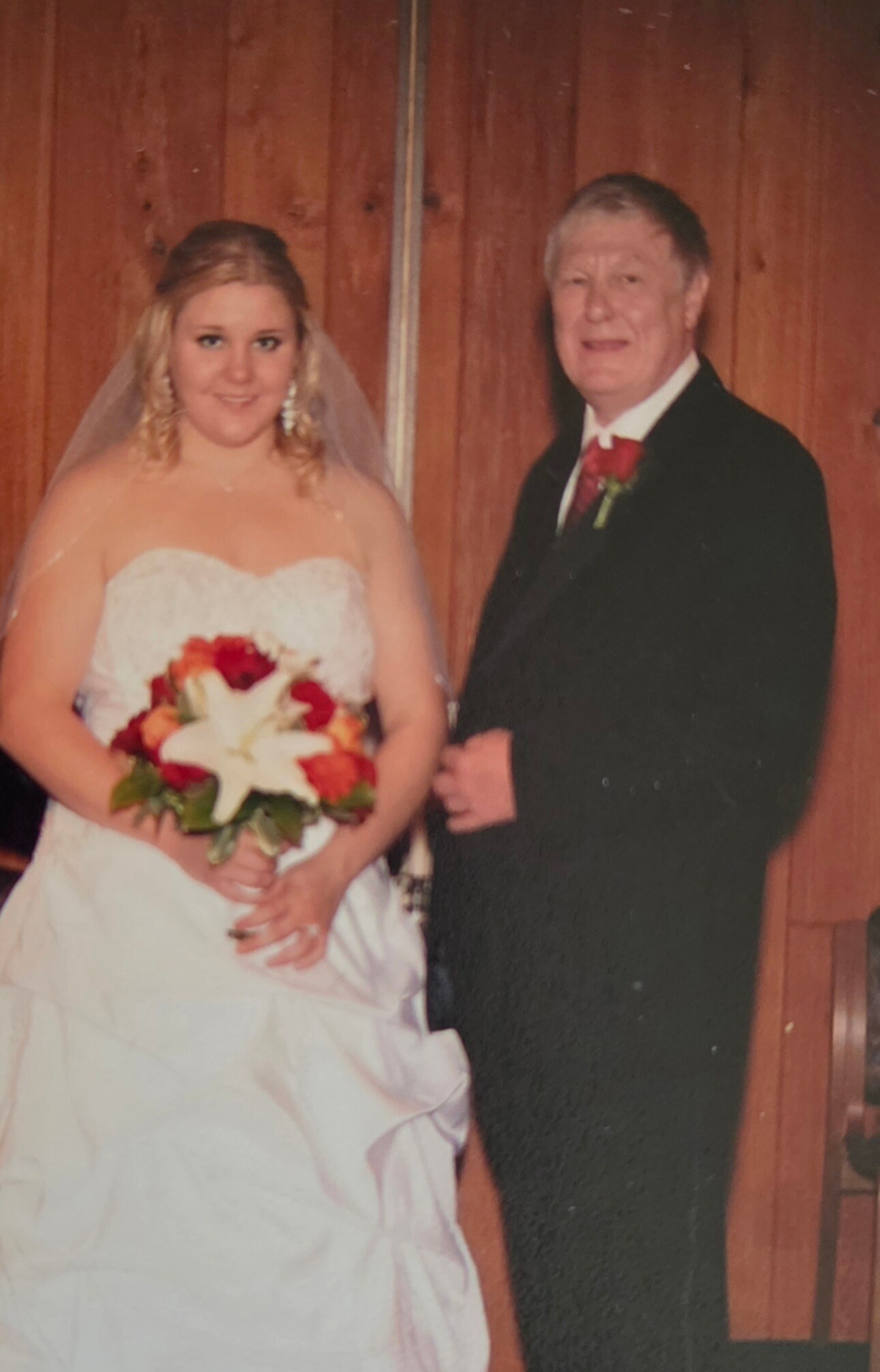 Bride in a white strapless wedding gown with a veil, holding a red-and-white bouquet, standing beside a man in a black suit with a red boutonniere against a wooden backdrop.