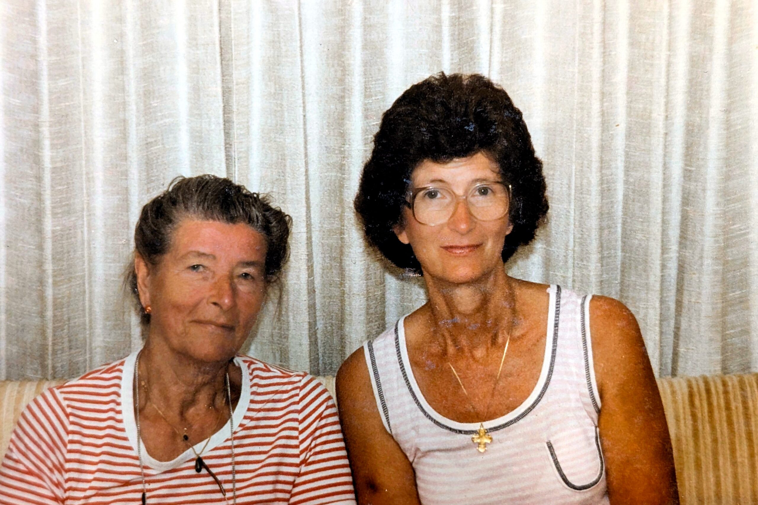 Two women sit side by side on a striped couch, wearing red-and-white and white-with-black-striped tops, posing for a photo in front of curtains.