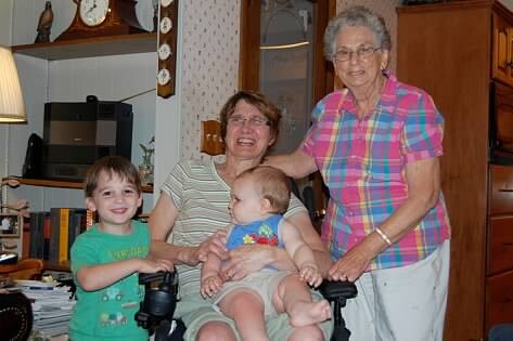 Three generations in a kitchen: a smiling boy in a wheelchair, a baby on a woman's lap, and an elderly woman standing beside them.