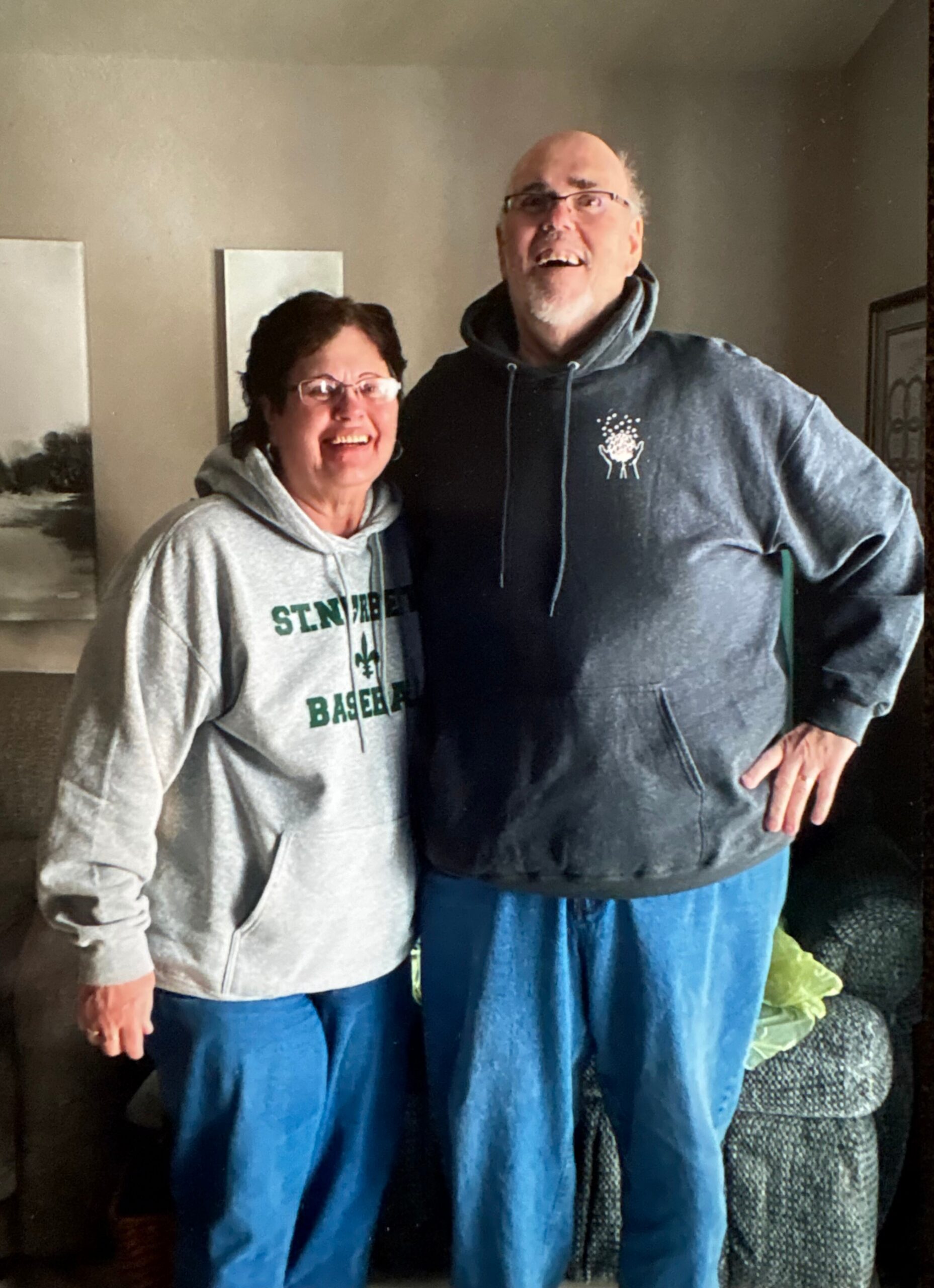 Two smiling adults stand side by side in a living room, wearing hoodies and blue jeans.