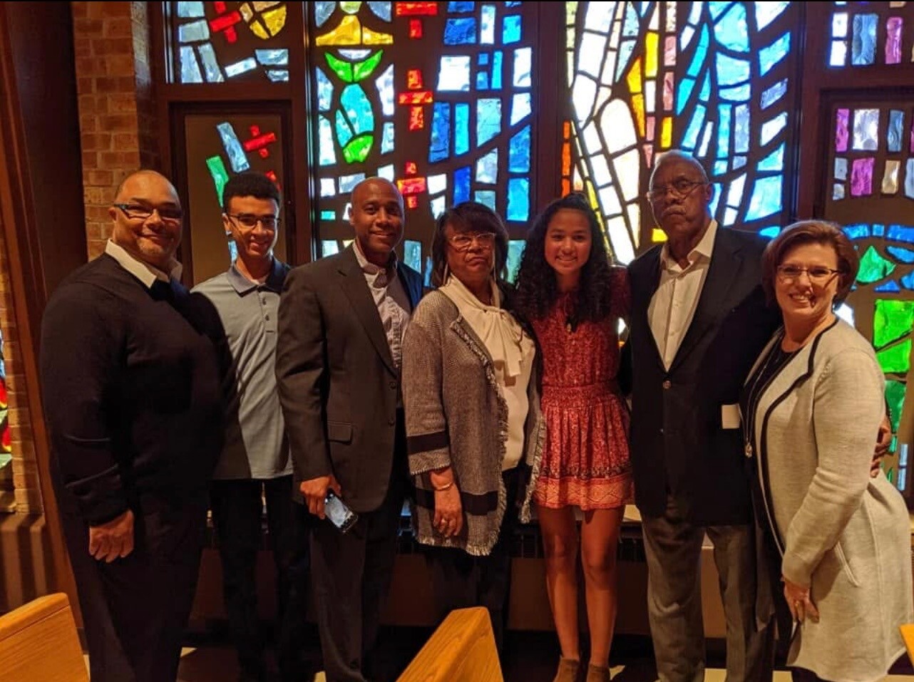 Eight adults stand shoulder to shoulder in front of a large colorful stained-glass window inside a building, smiling for the photo.