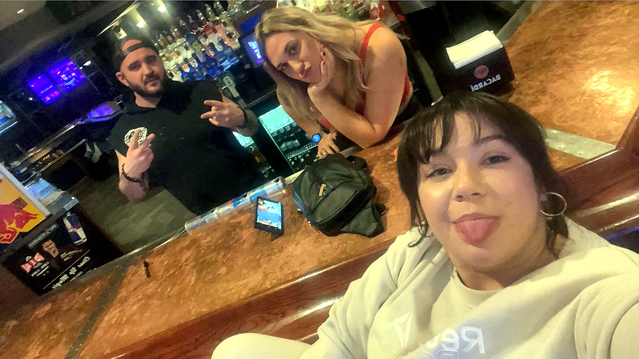 Close-up selfie of a woman in a white sweatshirt sticking out her tongue at a bar, with two friends and a liquor display in the background.