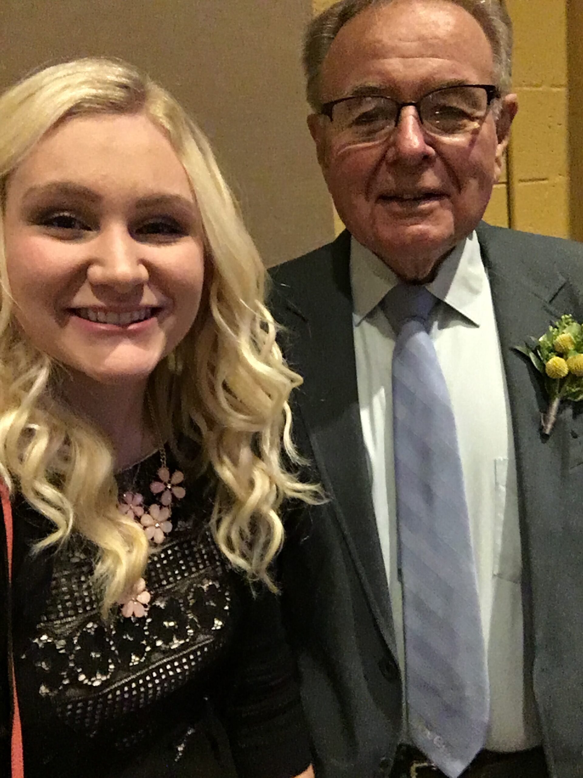 Smiling woman with long blonde hair and a floral necklace beside a man in a suit and tie at an indoor event.