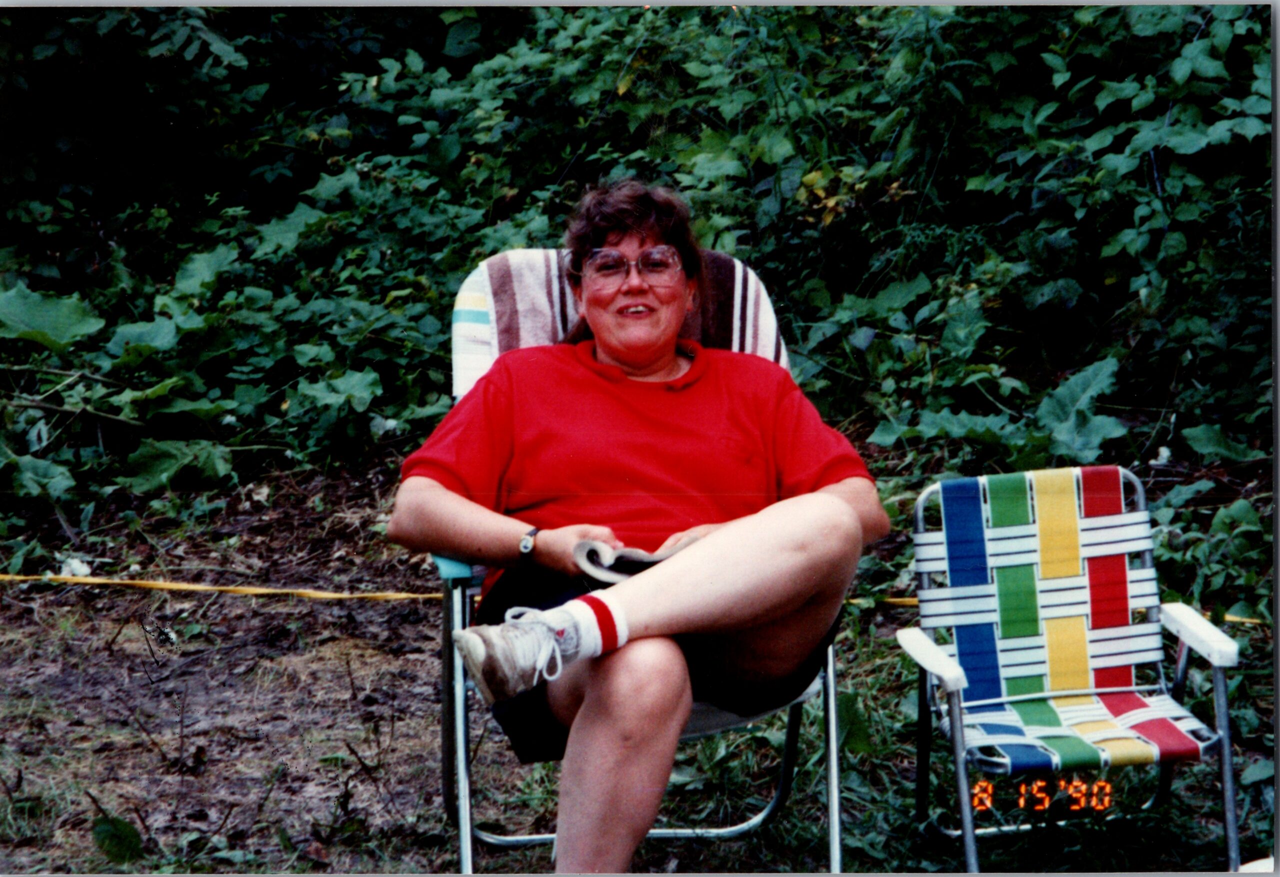 Smiling woman in a red shirt sits in a striped folding chair outdoors, surrounded by dense green foliage.