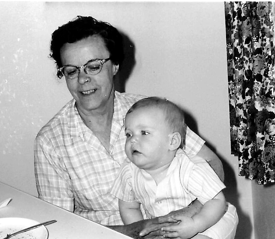 Smiling grandmother with glasses sits at a table, holding a baby on her lap.