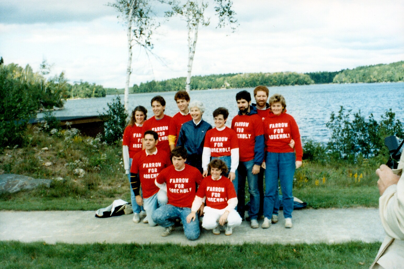Group of people wearing matching red shirts with 'FARROW FOR ASSEMBLY' posing together outdoors by a lake.