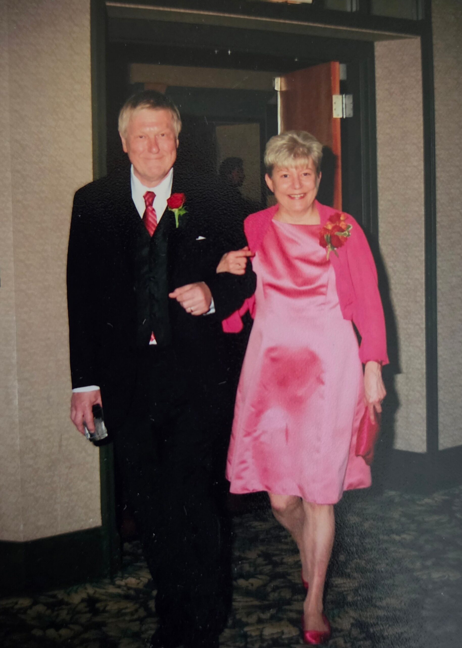Older couple posing together, man in a black suit with red tie and rose boutonniere, woman in a pink dress and matching jacket with a corsage, standing arm in arm in a doorway.