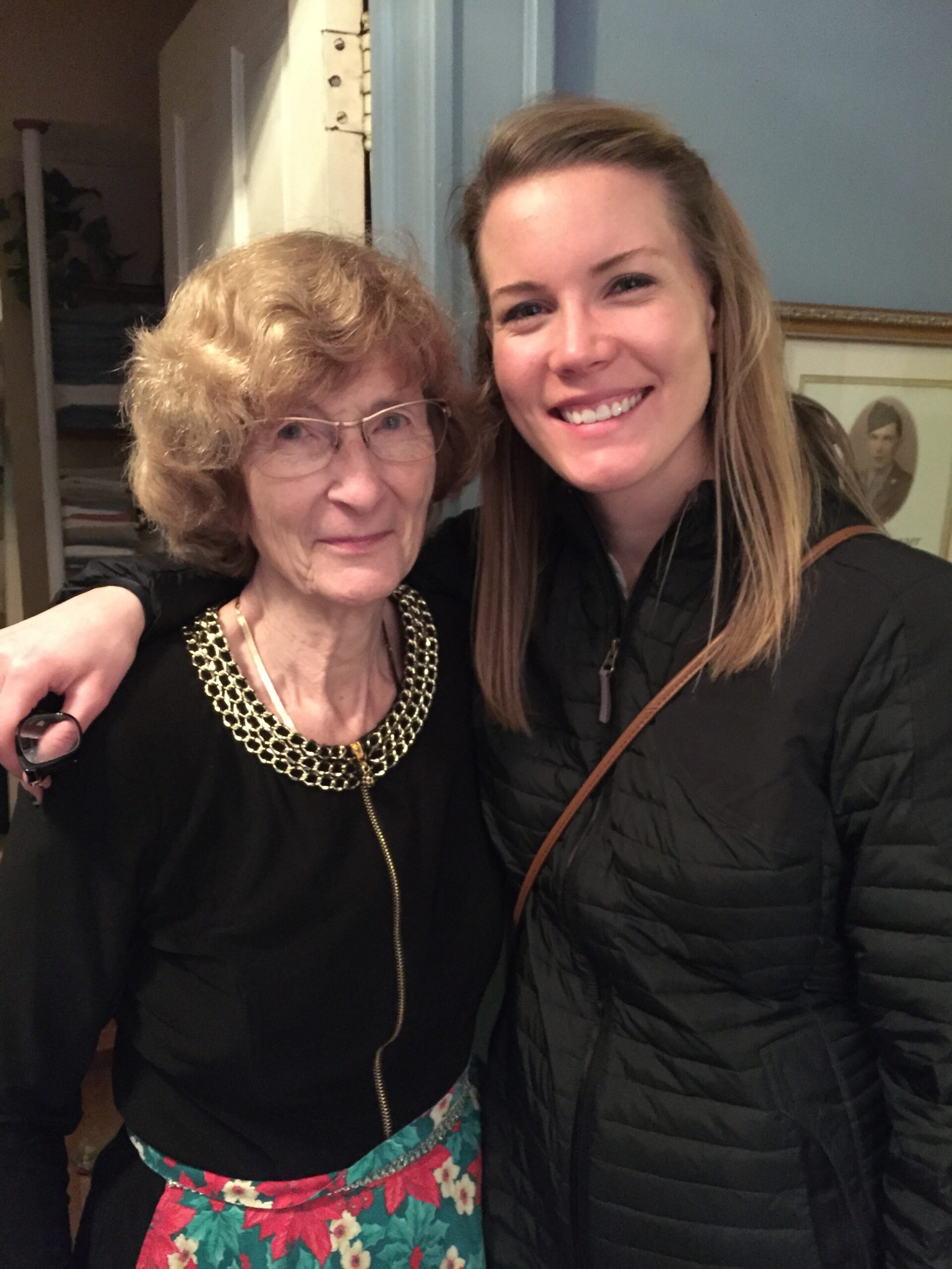 Two women smiling at the camera, an older woman with glasses and a gold necklace on the left and a younger woman in a black jacket on the right.
