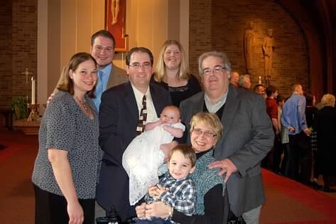 A multi-generational family posing inside a church, holding a baby in a white blanket, with brick walls and religious statues in the background.