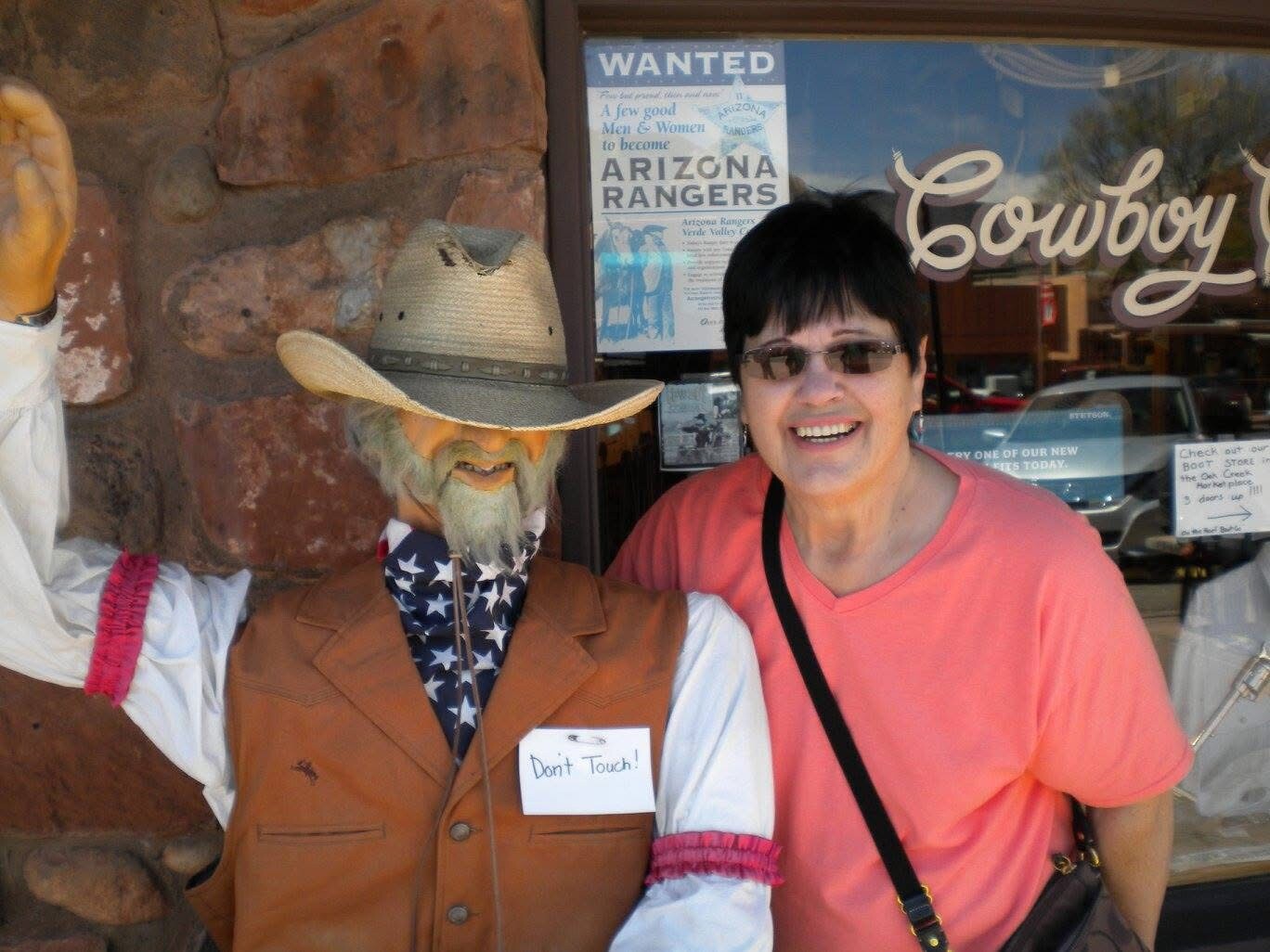 Smiling woman in a pink shirt posing with a life-size cowboy mannequin wearing a hat and a 'Don't Touch!' tag outside a shop window.