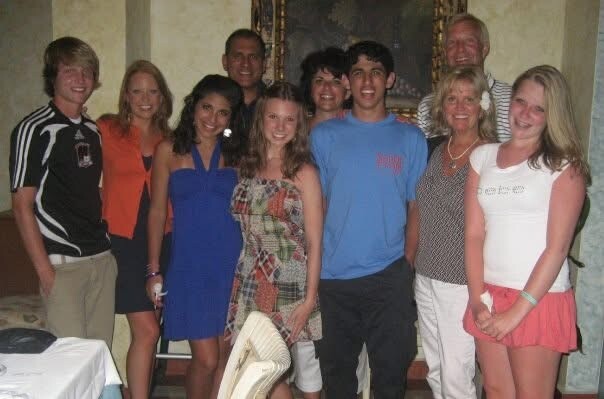 Group of smiling young adults posing together indoors at a casual event. Everyone stands close, some wearing bright outfits.