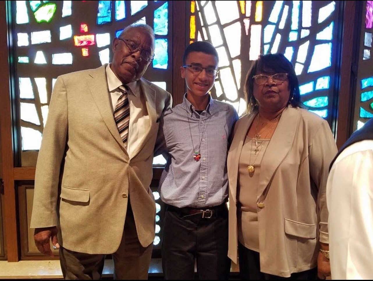 Three people posing indoors in front of a colorful stained-glass window: an older man in a light suit, a boy in a blue check shirt, and a woman in a beige blazer.