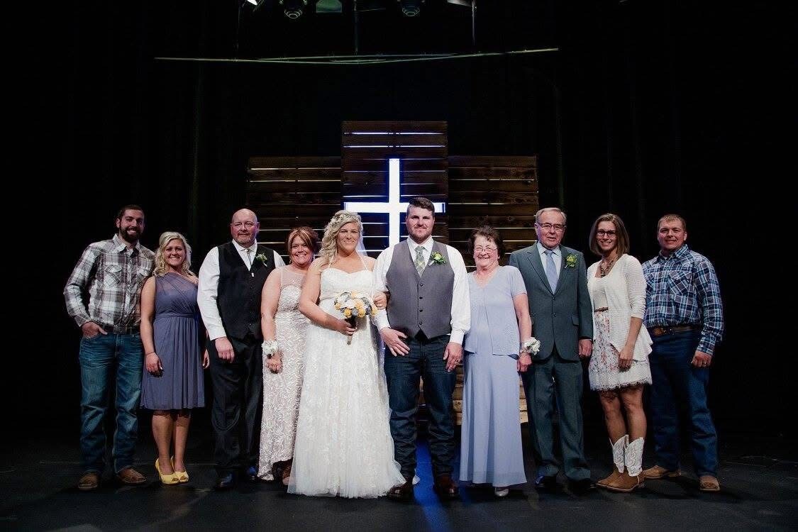 Bride and groom pose with their wedding party on a stage, smiling, with a wooden cross backdrop behind them.