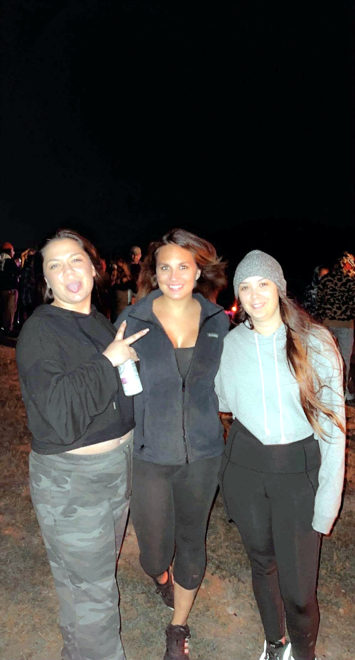 Three women posing together at night outdoors, smiling for the camera; one holds a bottle and peace sign on the left, all in athletic wear.