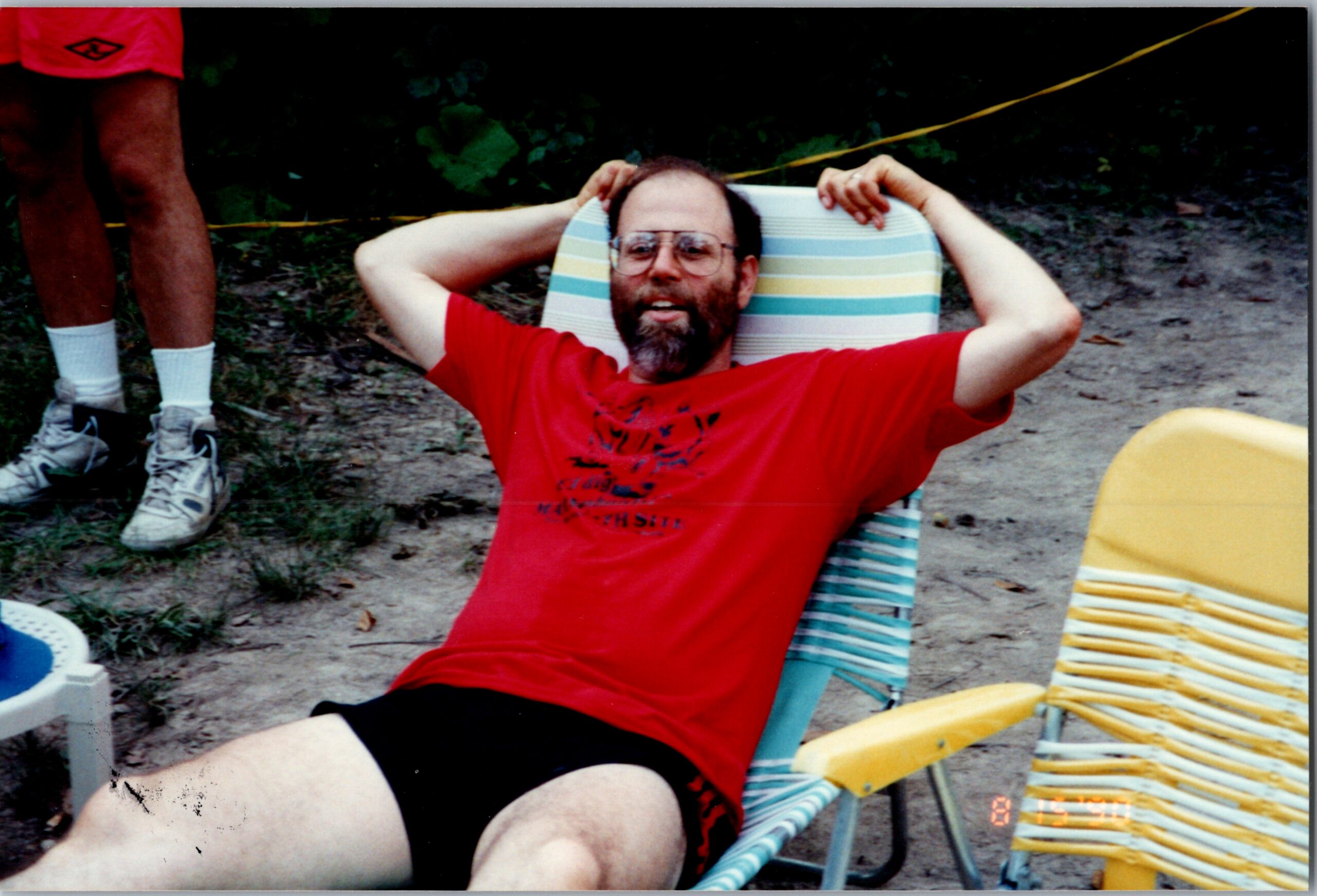 Man with a beard and glasses relaxing in a striped beach chair wearing a red shirt, outdoors on a sandy area.