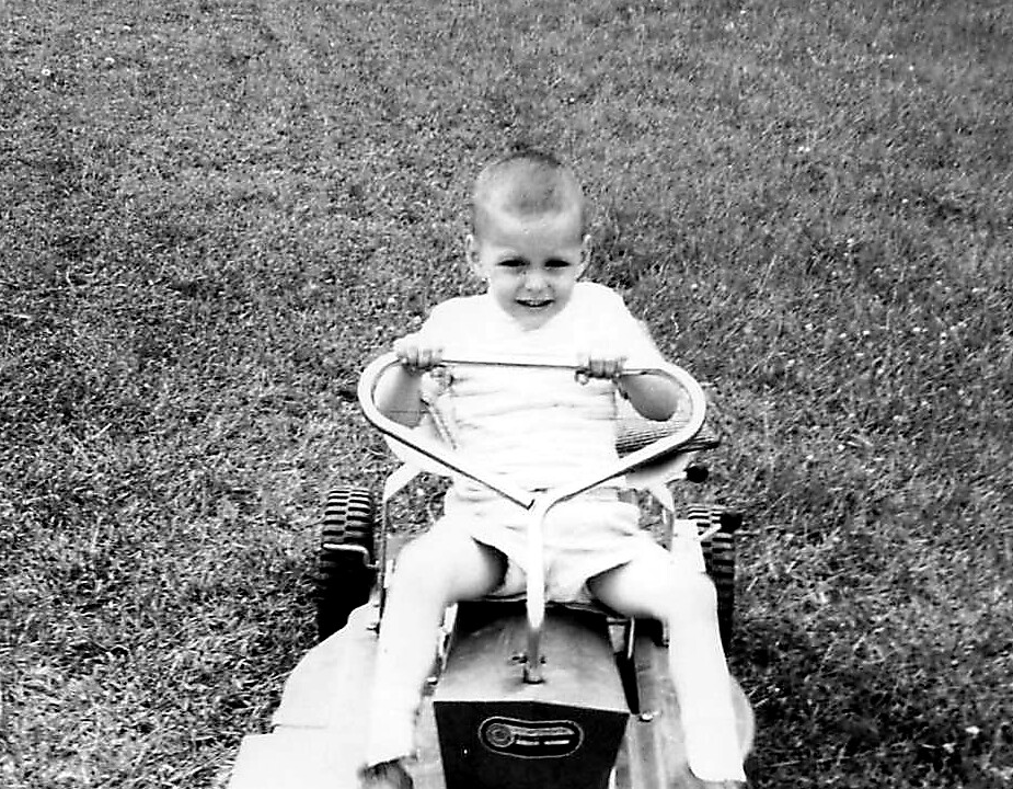 Toddler smiling while riding a small tricycle on a grassy lawn, black-and-white photo.