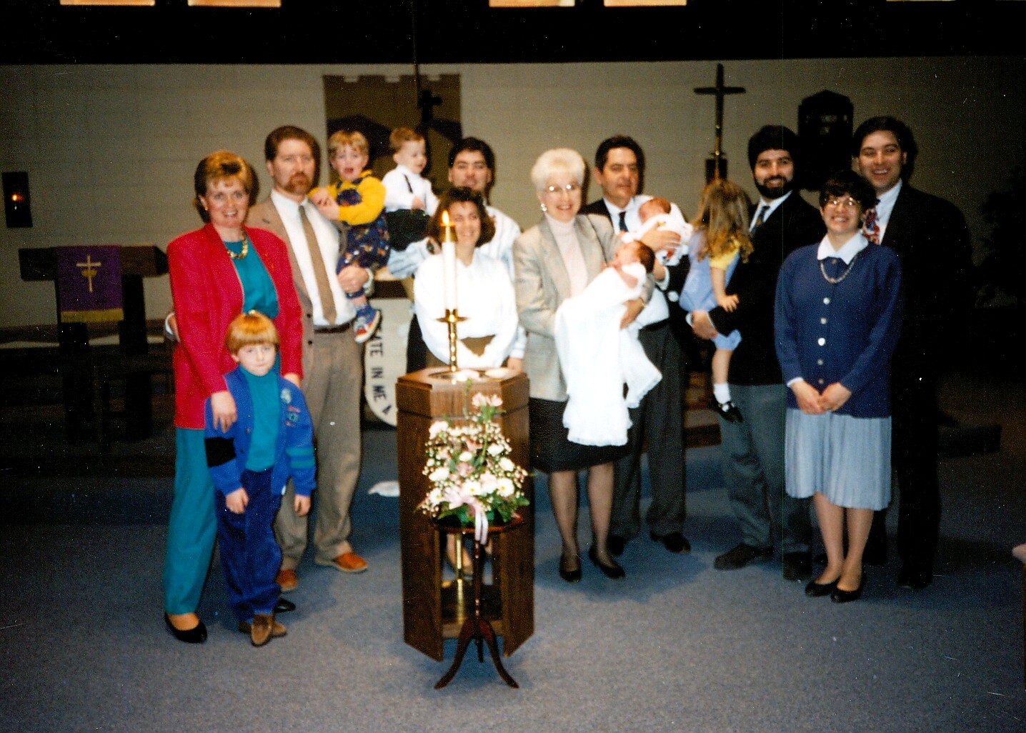Family and church members pose for a group photo in a sanctuary around a baptismal stand with a lit candle and flowers in front of a cross backdrop.