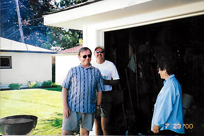 Two men wearing sunglasses stand in a sunlit backyard near an open garage while a woman in a blue shirt faces them.