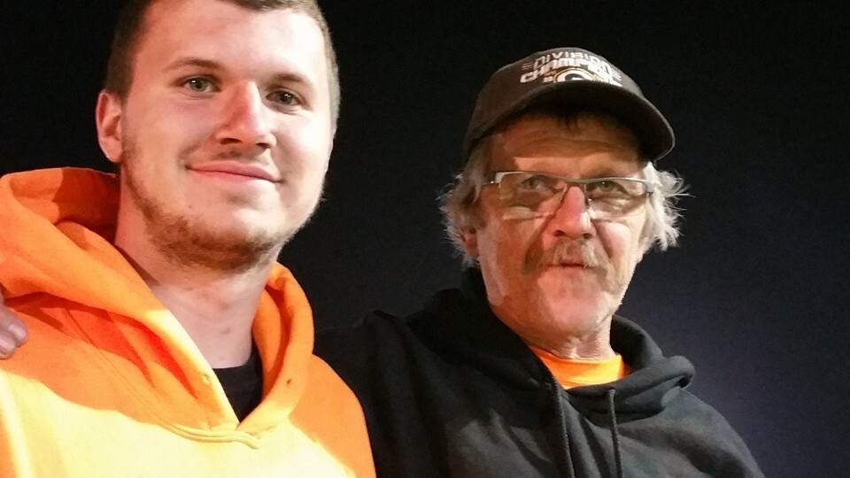 Two men posing for a selfie: a younger man in an orange hoodie and an older man in a black hoodie and baseball cap, both smiling against a dark background.