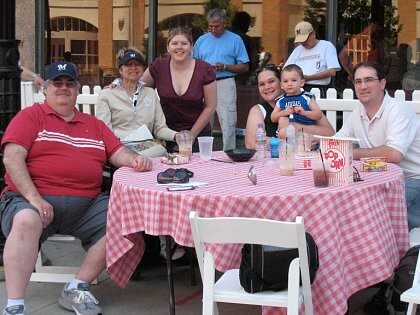 Group of eight people around a pink-checkered table at an outdoor café; a child sits with a man on the right among drinks and snacks.