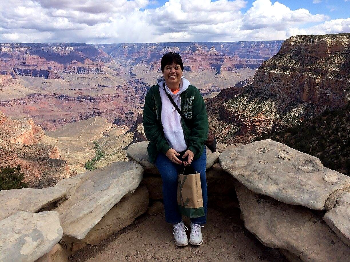 Woman sits on rock ledge at the Grand Canyon, smiling, wearing a green jacket and white hoodie, holding a paper bag.